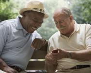 2 older men sit on a park bench. One is showing a phone screen to the other. They are both smiling broadly.
